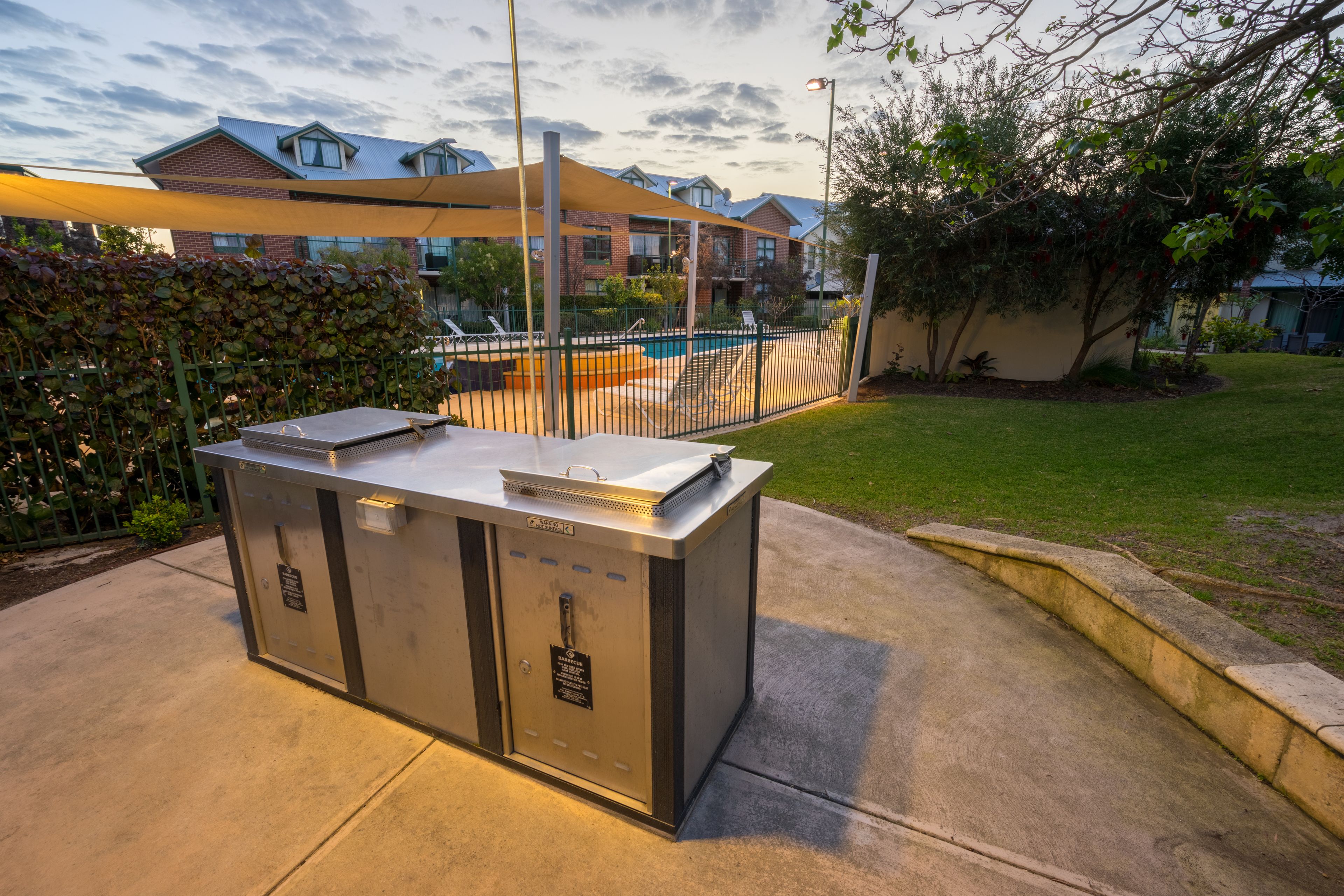 Een draagbare grillstation met een metalen ombouw, op een betonnen terras met een zwembad en een hek op de achtergrond.