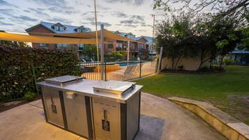 Een draagbare grillstation met een metalen ombouw, op een betonnen terras met een zwembad en een hek op de achtergrond.