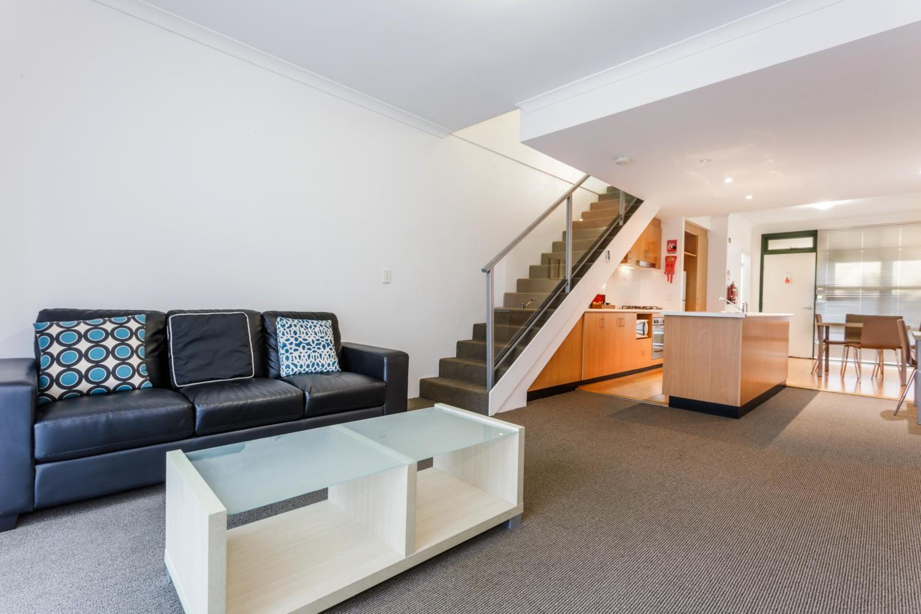A modern living room with a black leather sofa, a glass coffee table, and a staircase leading to a kitchen area.