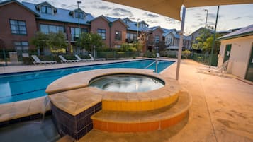 Una piscina con un jacuzzi, rodeada de sillas de descanso y un edificio de ladrillo al fondo.