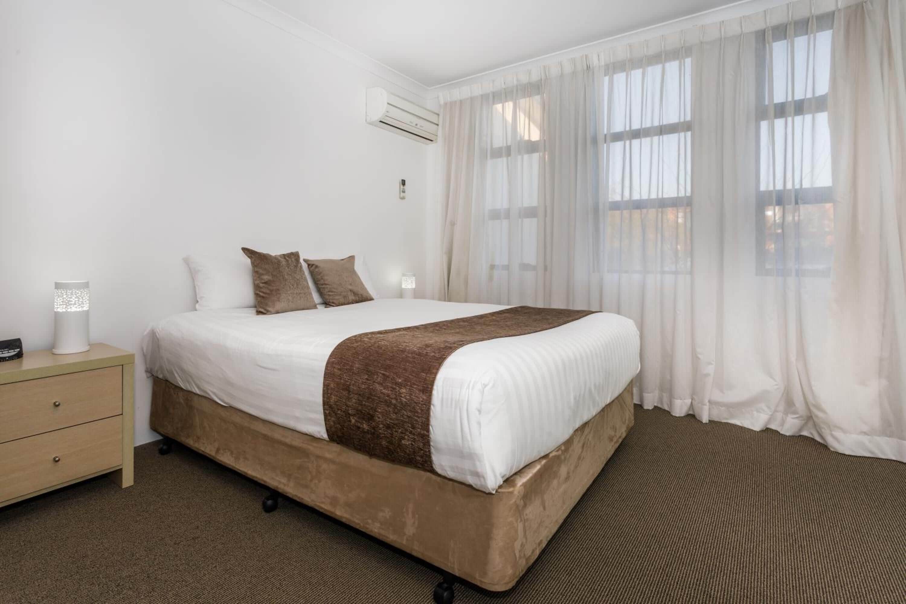 A hotel room with a bed, a nightstand, a lamp, and a window with sheer curtains.
