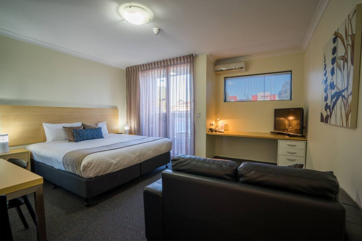 A hotel room with a bed, a desk, a sofa, a TV, and a window with blinds.