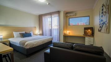 A hotel room with a bed, a desk, a sofa, a TV, and a window with blinds.
