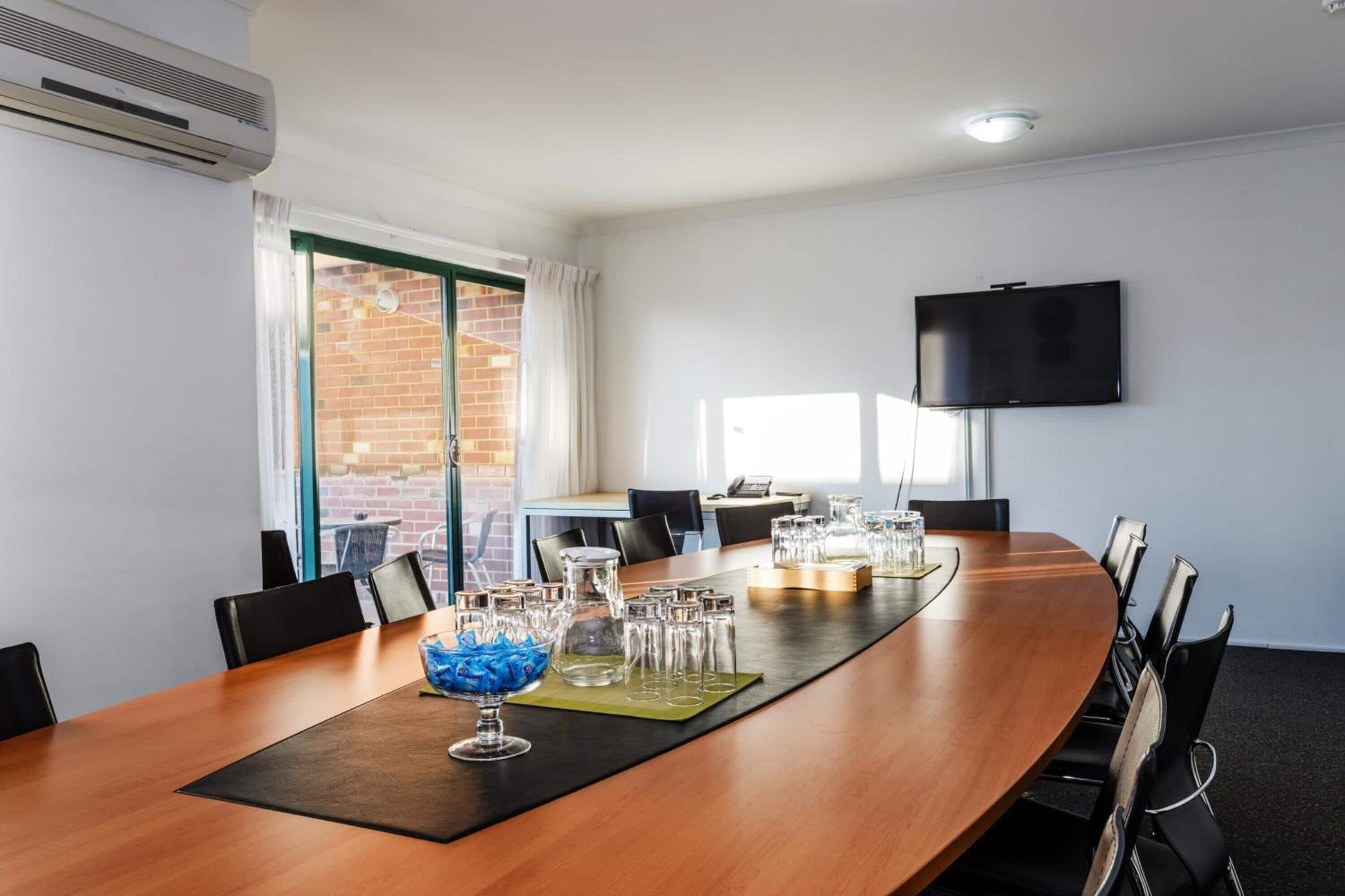 A conference room with a long wooden table, chairs, a TV mounted on the wall, and a window with curtains.