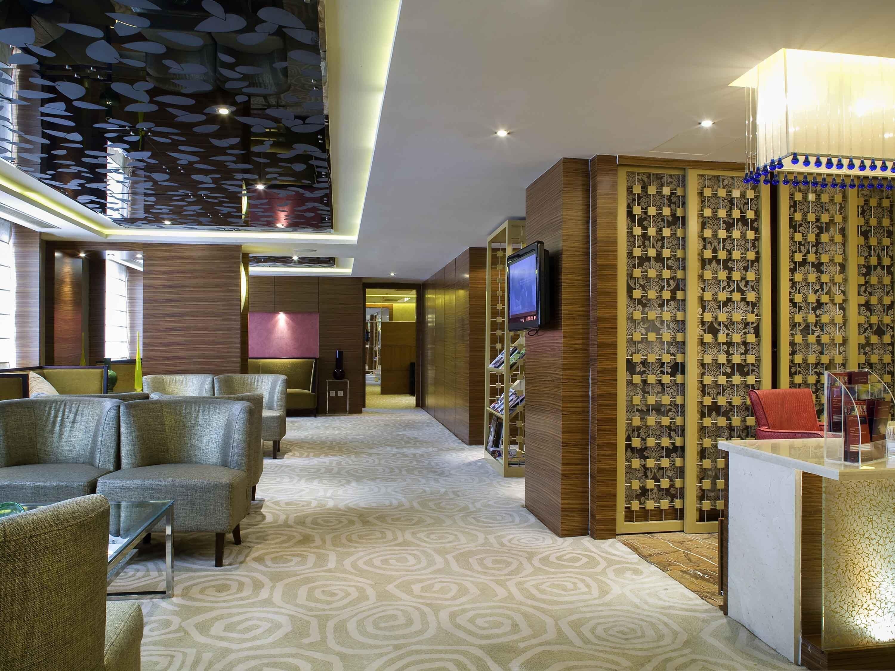 Eine moderne Hotelhalle mit gemustertem Teppich, einer Sitzecke mit Sofas, einem Fernseher auf einem Holzständer und einer Wand mit geometrischem Dekor.