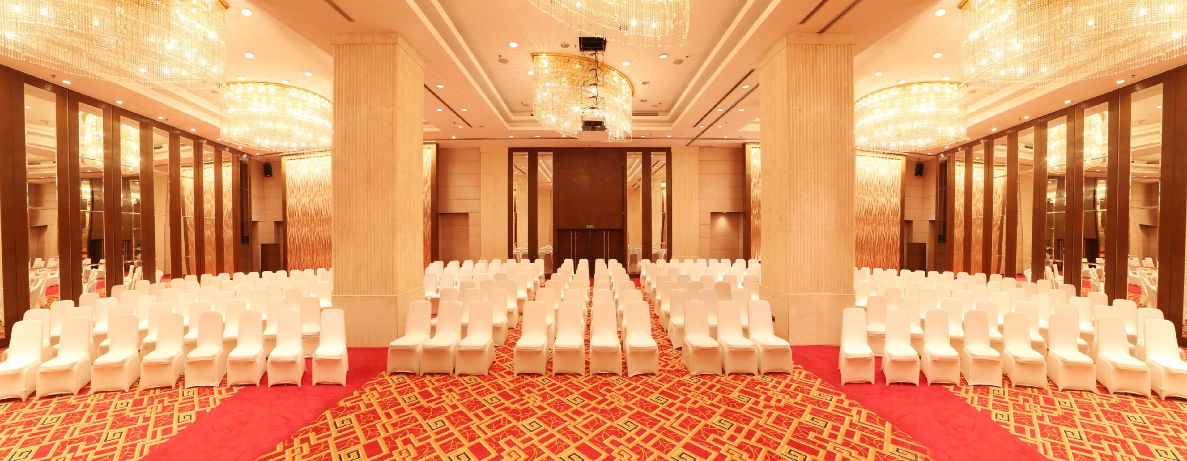 A spacious conference room with rows of white chairs, a red carpet, and ornate chandeliers.