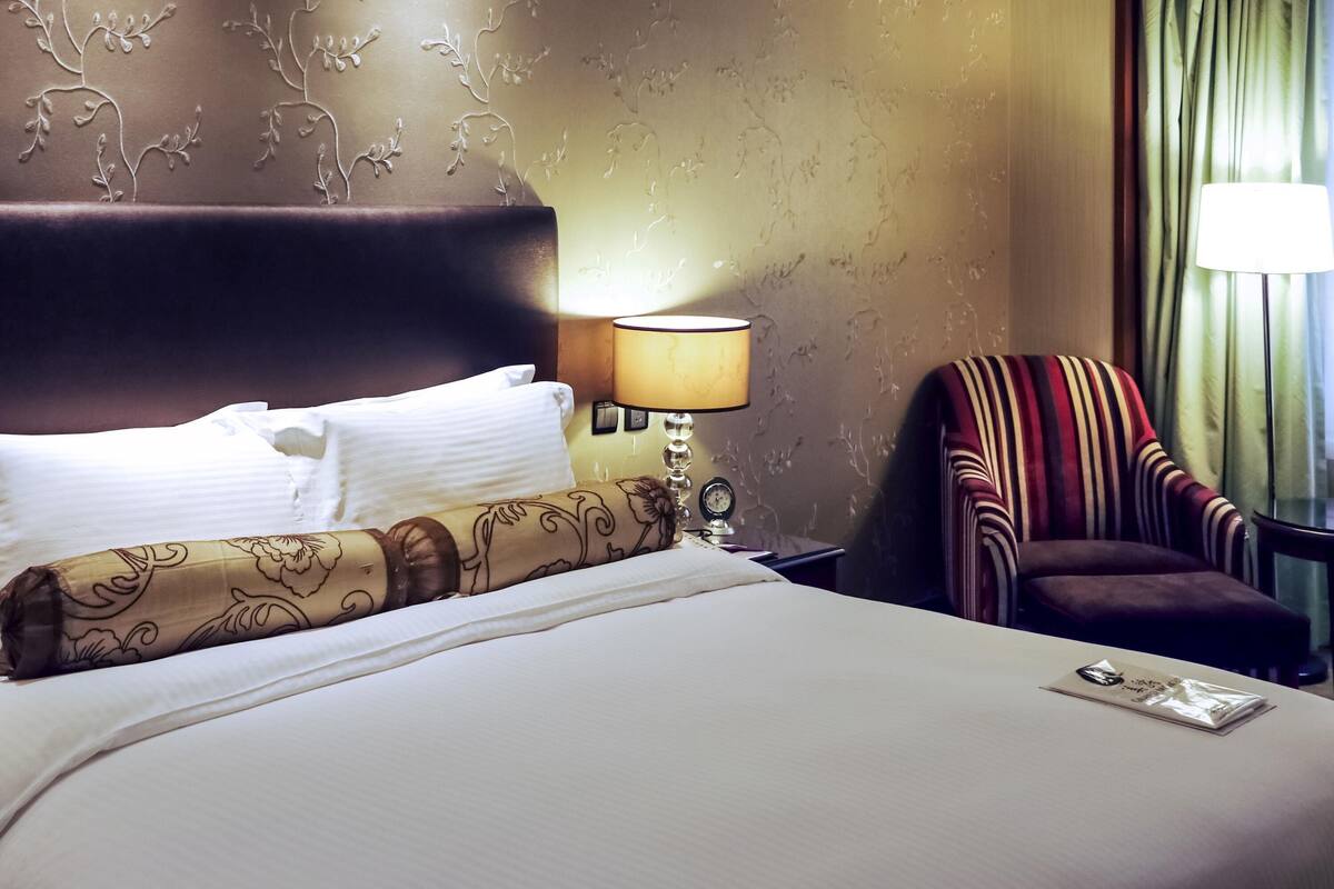 Een hotelkamer met een bed, een nachtkastje met een lamp, een stoel en een raam met gordijnen.