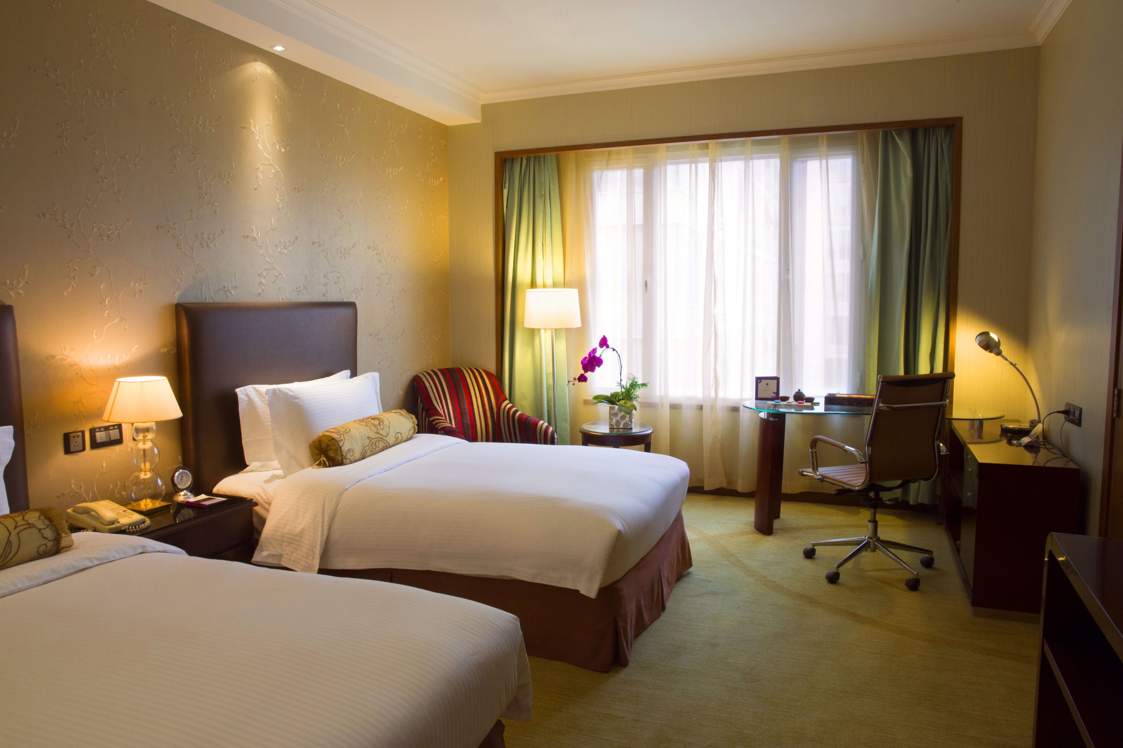 A hotel room with two beds, a desk, a chair, a lamp, and a window with curtains.