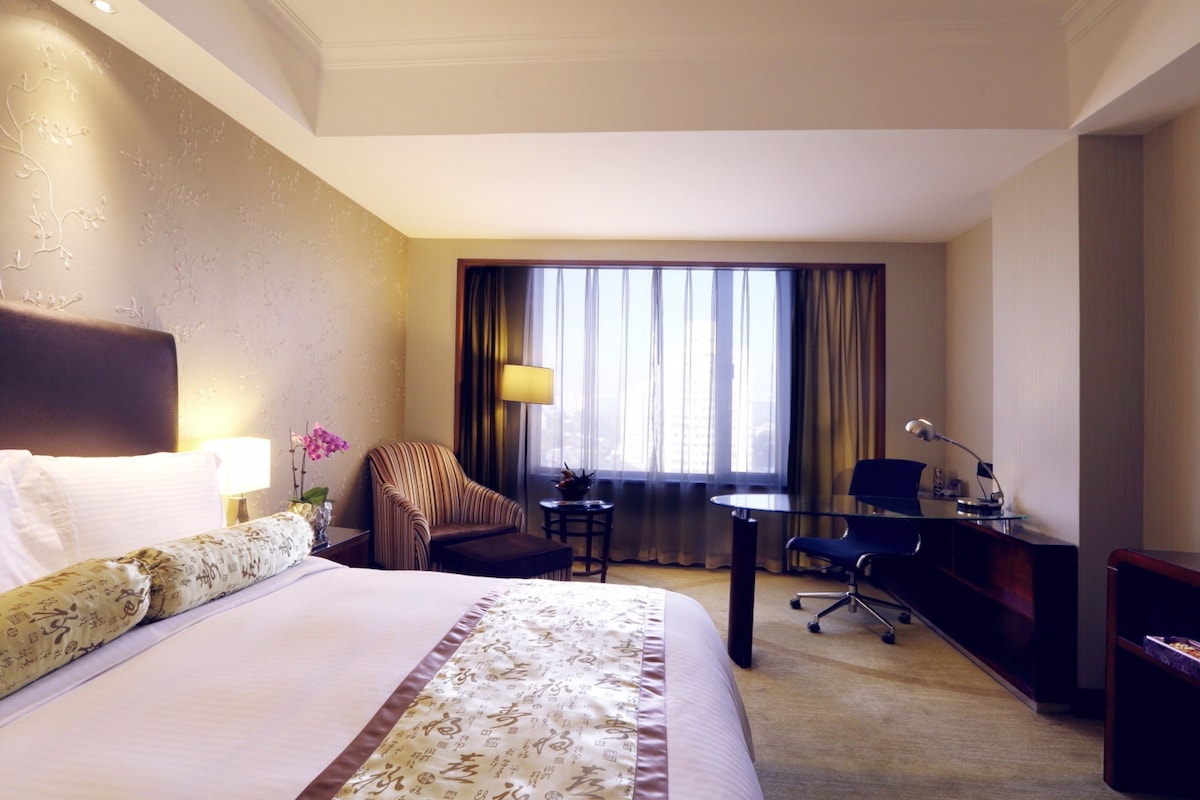 Habitación de hotel con una cama grande, un escritorio, una silla, una lámpara y un ventanal con cortinas.