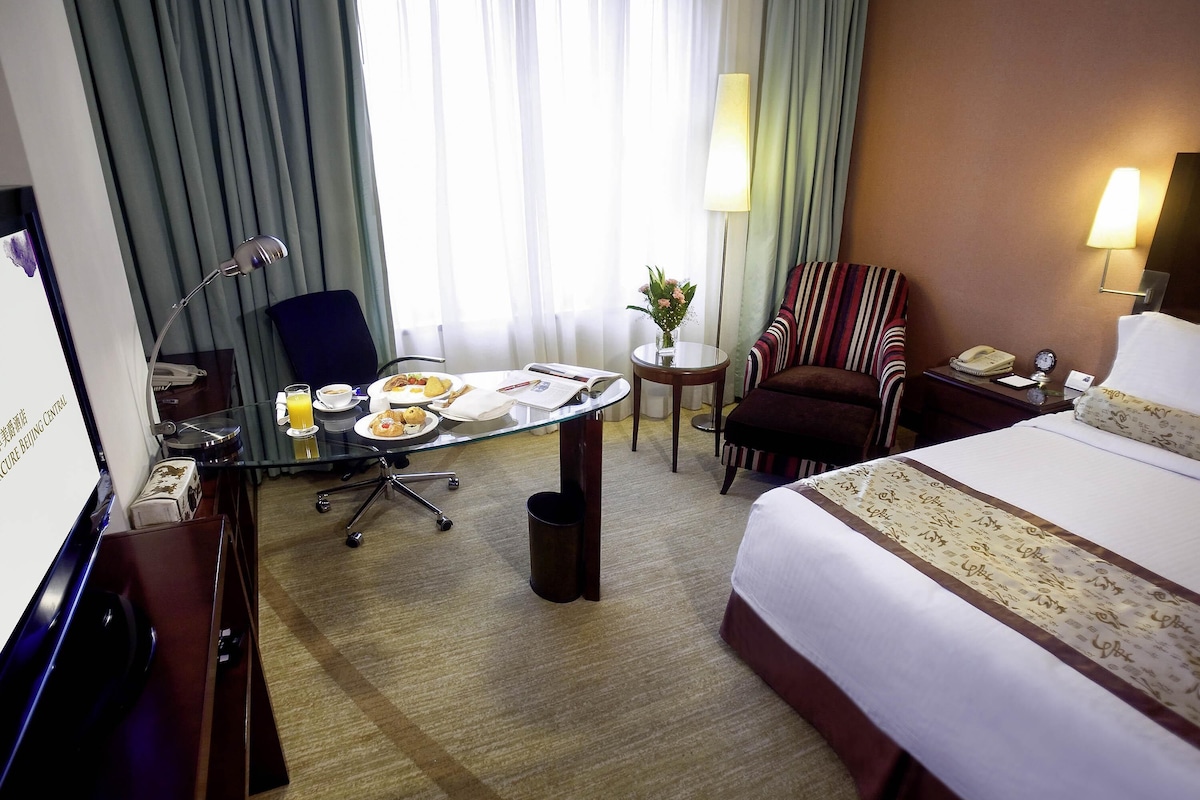Una habitación de hotel con una cama, un escritorio con lámpara, una silla, una mesita con un jarrón y una ventana con cortinas.