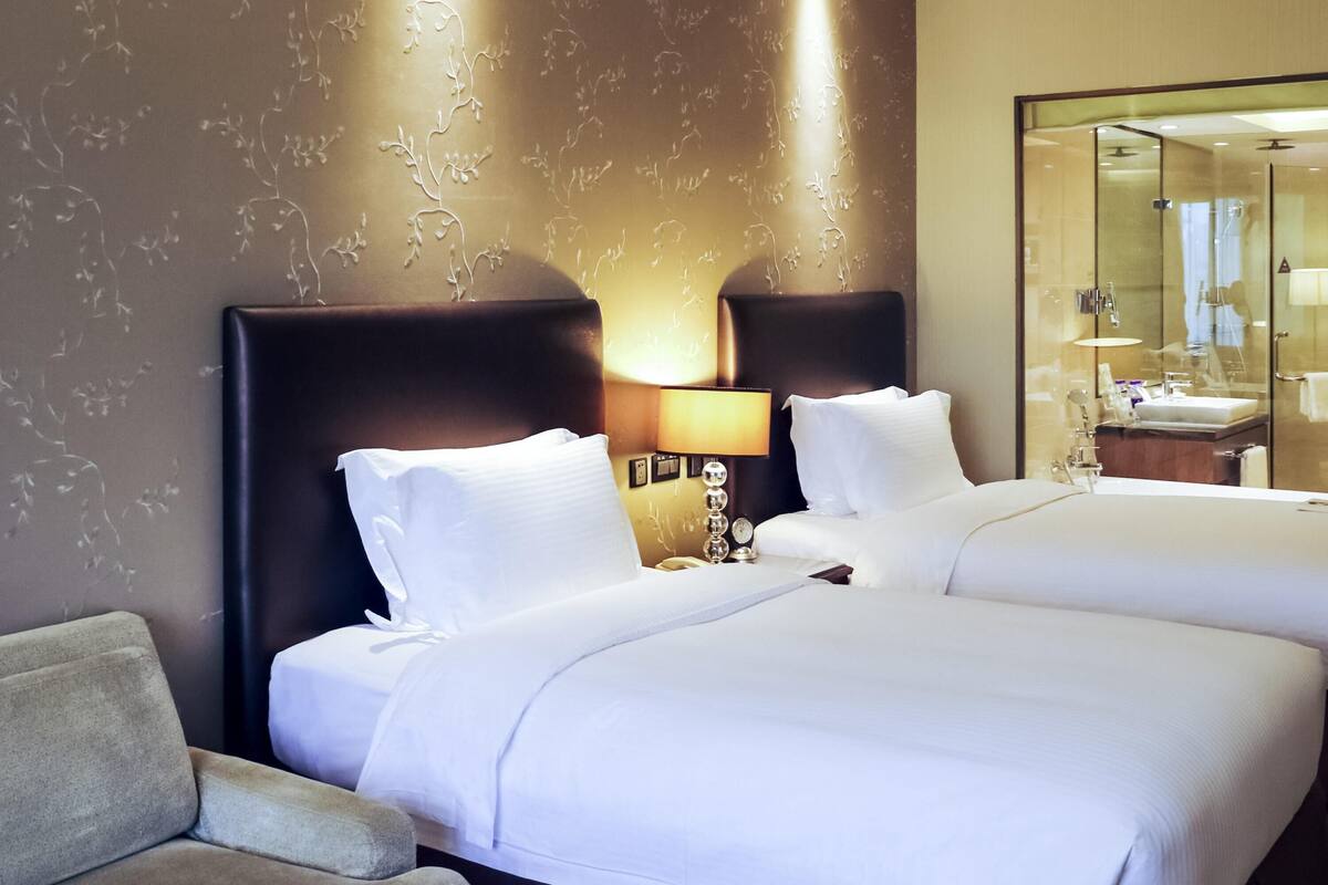 Een hotelkamer met twee bedden, een bank, een nachtkastje met lamp en een zichtbare badkamer via een glazen deur.