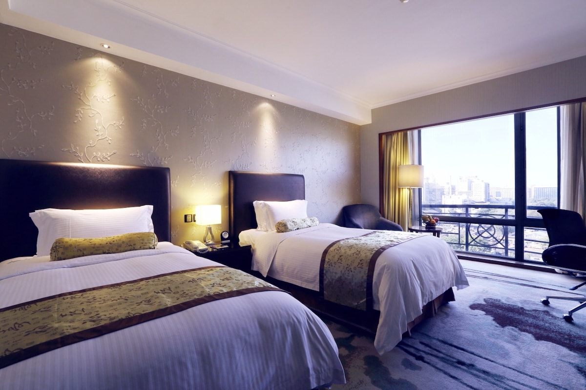 Habitación de hotel con dos camas, un ventanal grande con vistas a la ciudad, un escritorio con lámpara y una silla.
