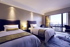 Habitación de hotel con dos camas, un ventanal grande con vistas a la ciudad, un escritorio con lámpara y una silla.