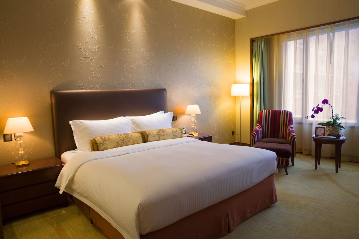 Una habitación de hotel con una cama grande, dos mesitas de noche con lámparas, un sillón a rayas, una pequeña mesa con una planta y una ventana con cortinas.