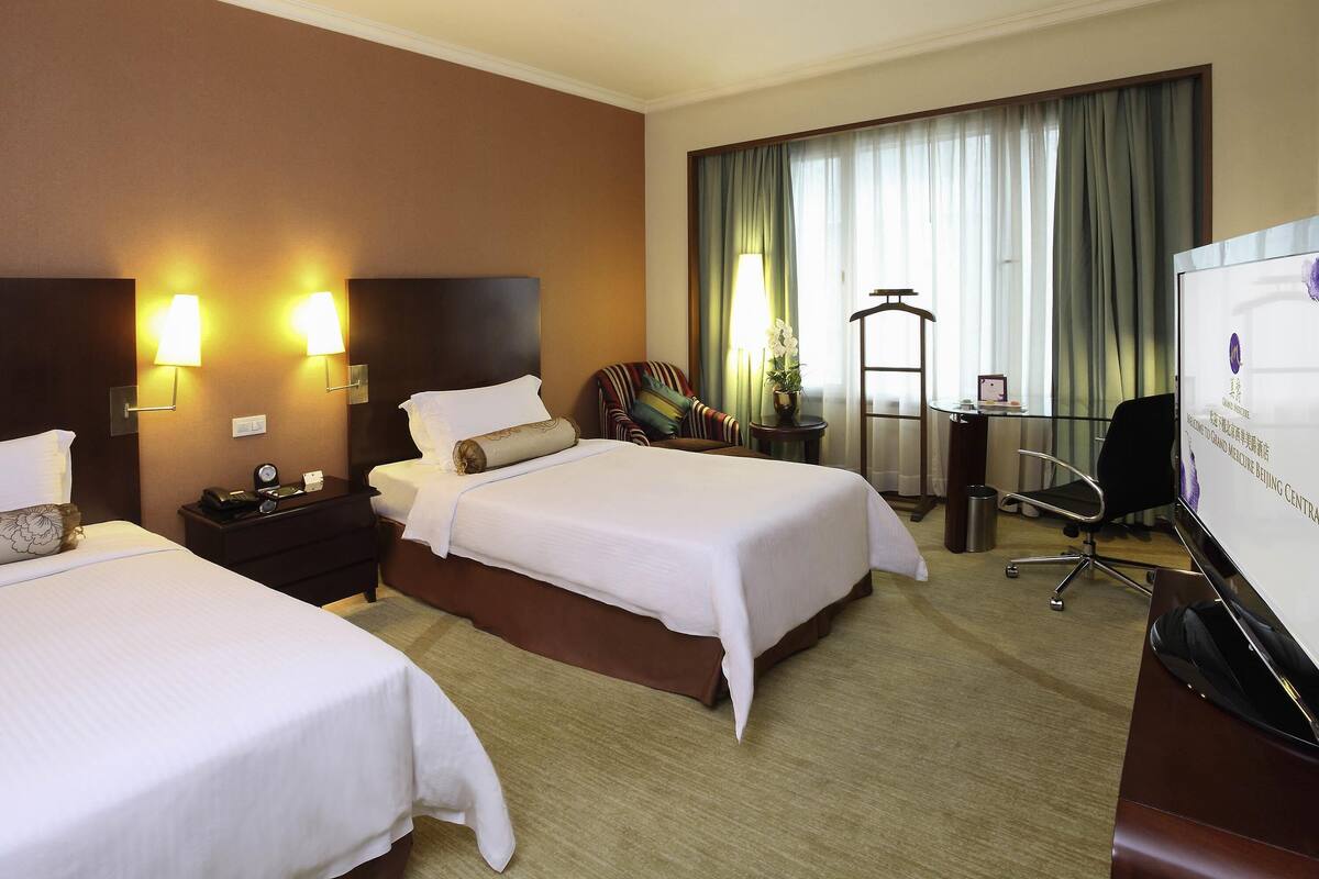 Een hotelkamer met twee bedden, een bureau, een stoel en een groot raam met gordijnen.
