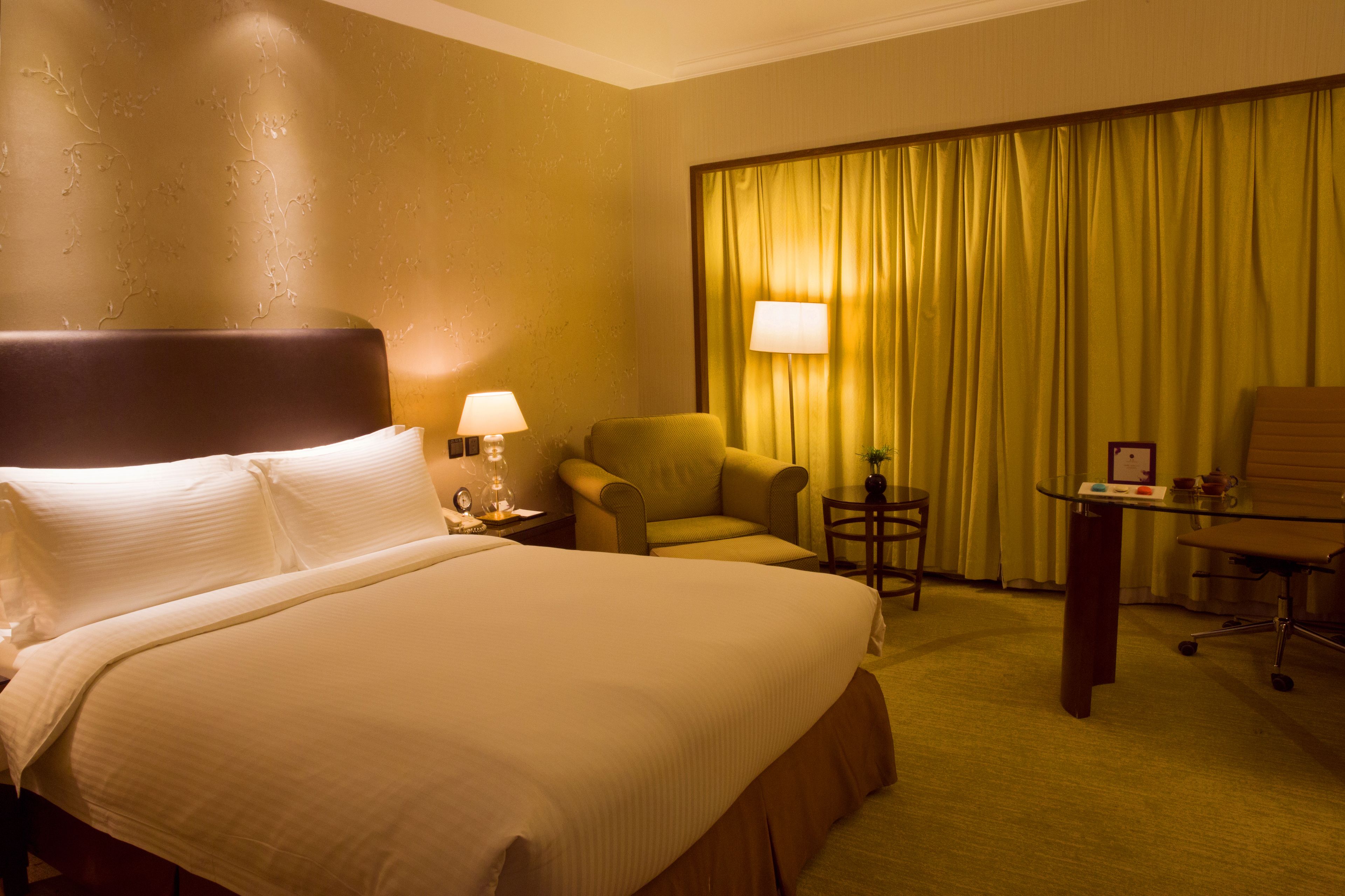 Quarto de hotel com uma cama grande, duas cadeiras, uma mesinha e uma escrivaninha com abajur.