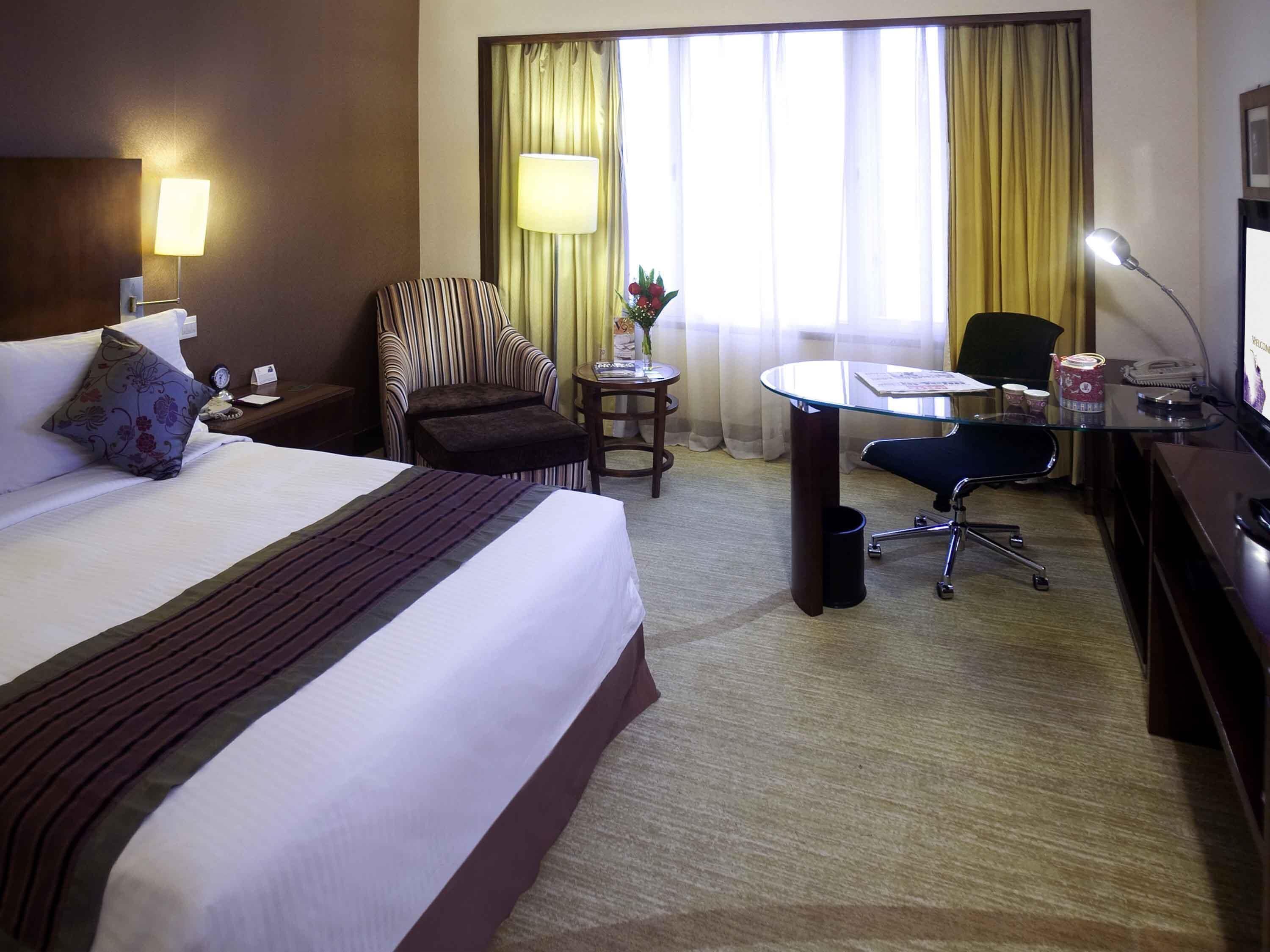 Een hotelkamer met een bed, een bureau, een stoel, een lamp, een klein tafeltje met een vaas en gordijnen.