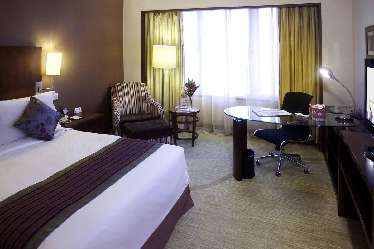 Een hotelkamer met een bed, een bureau, een stoel, een lamp, een klein tafeltje met een vaas en gordijnen.