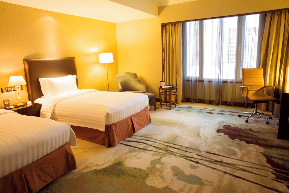 Habitación de hotel con dos camas, un escritorio, una silla y un ventanal con cortinas.