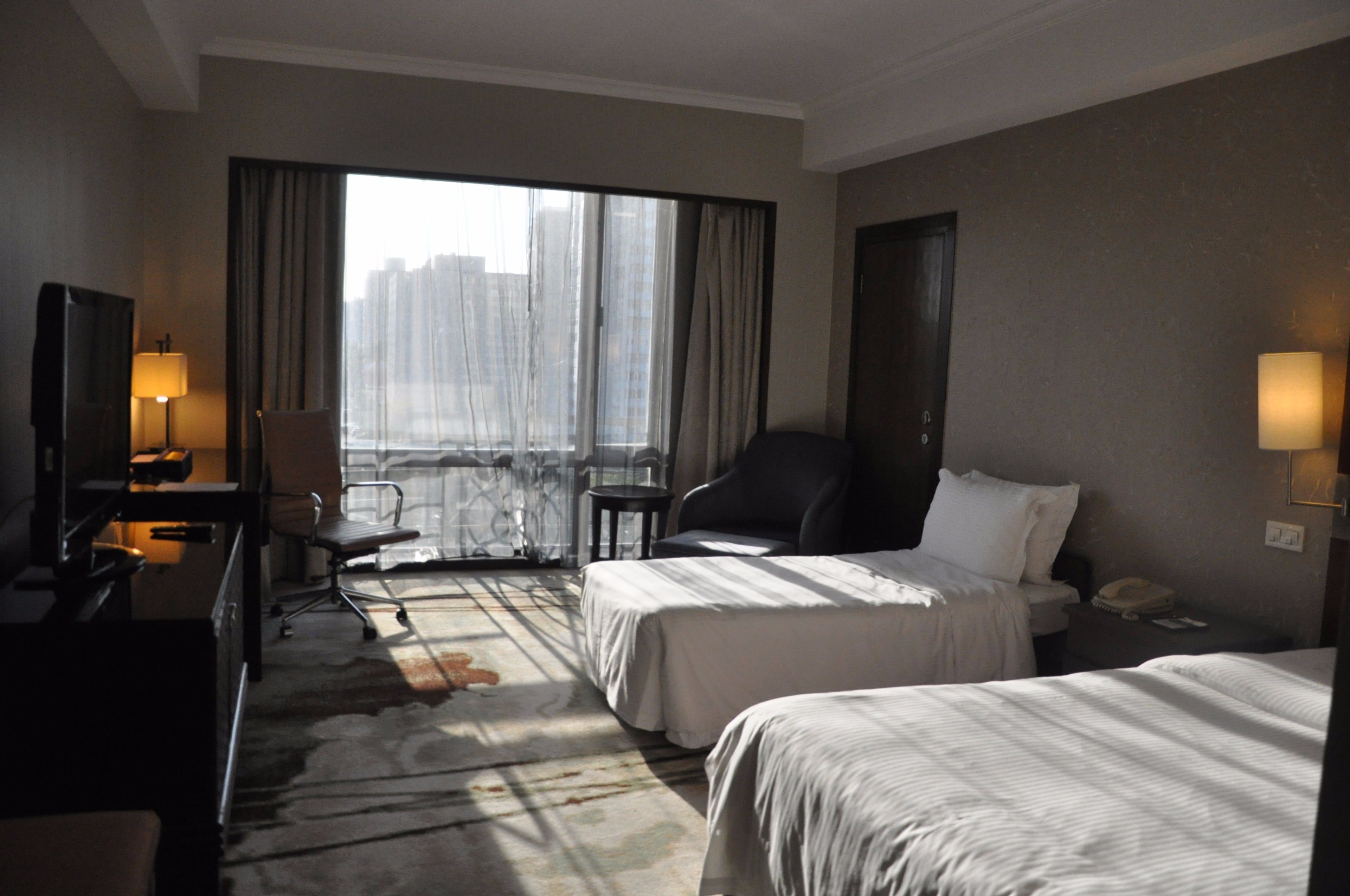 A hotel room with two beds, a desk with a lamp, a chair, and a view of a cityscape through large windows.