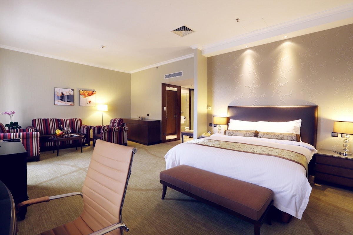 Una habitación de hotel con una cama grande, un espacio de descanso con sofá y sillones, un escritorio y una mesita con lámpara.
