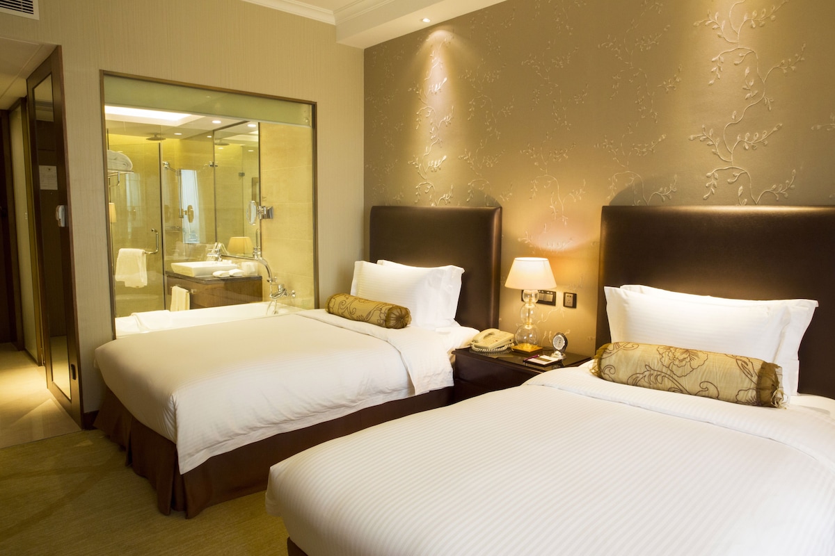 Habitación de hotel con dos camas, un espejo grande, baño con lavabo e inodoro, y mesita de noche con lámpara y teléfono.