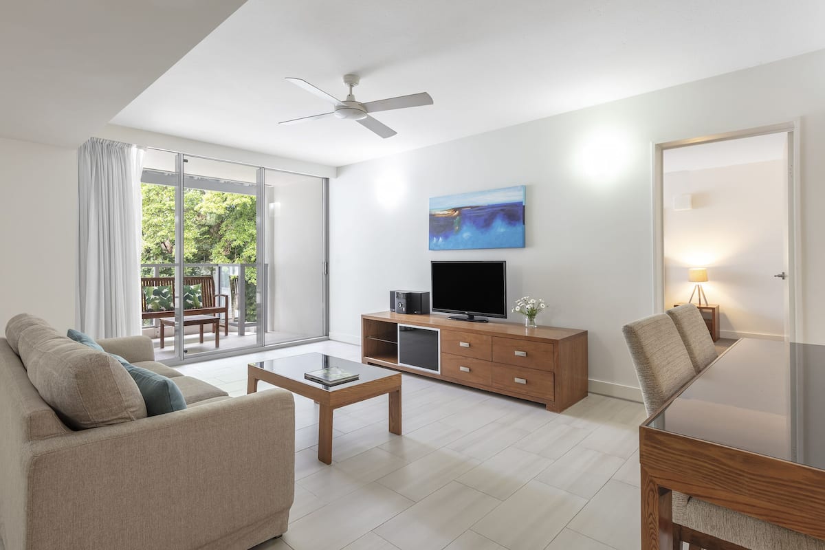 A modern living room with a sofa, coffee table, TV, and a balcony with a view of greenery.