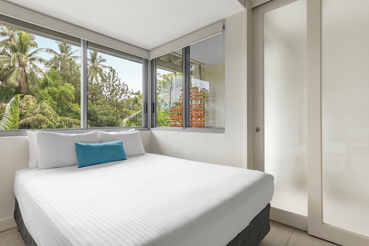 A modern bedroom with a large bed, white bedding, and a view of palm trees through large windows.
