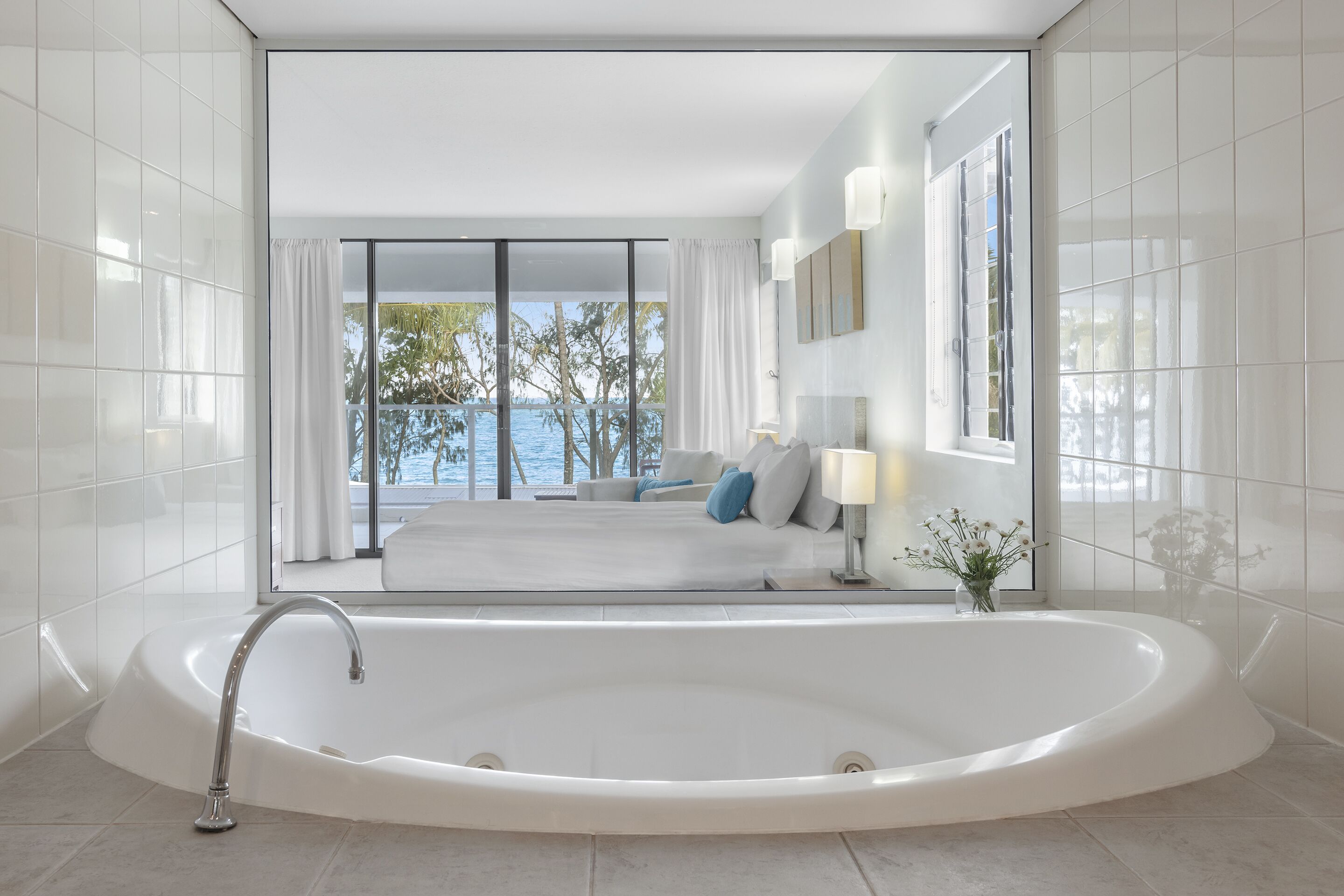 A modern bathroom with a large bathtub, a glass-enclosed shower area, and a view of a pool and palm trees.