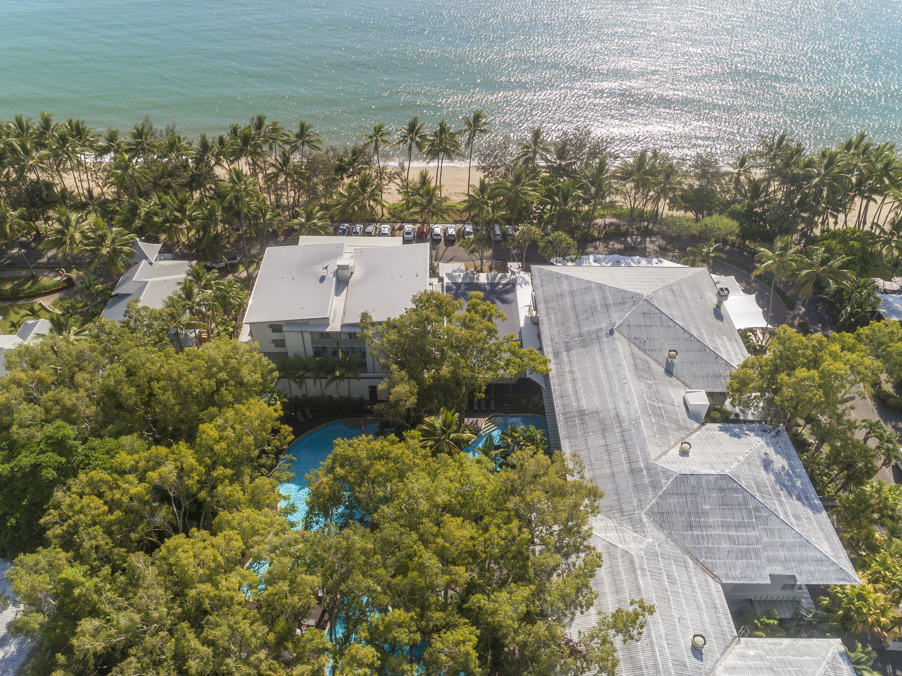 An aerial view of a resort with multiple buildings, a swimming pool, and a beach with palm trees.