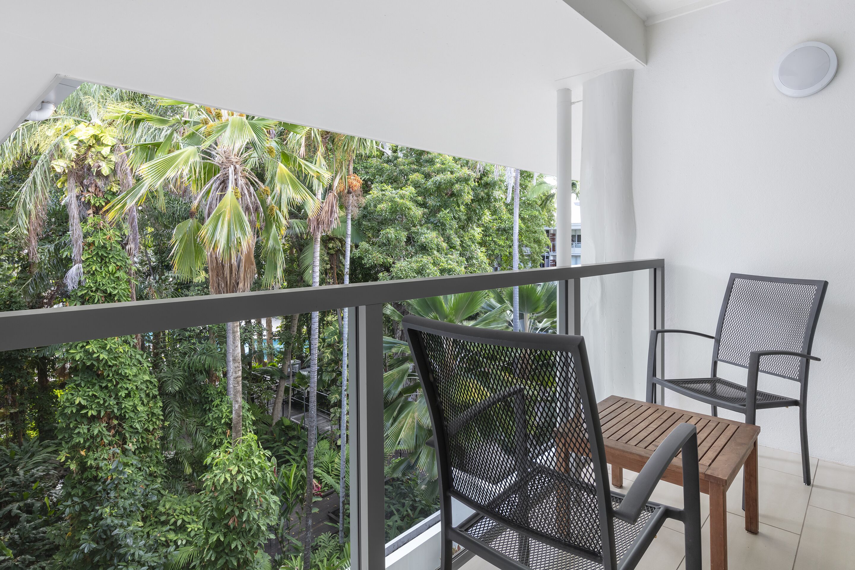 Un balcone con vista su una fitta vegetazione tropicale, un tavolo di legno e due sedie in metallo.