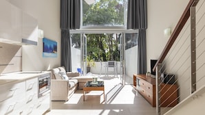 A modern living room with a sofa, coffee table, and TV. There is a balcony with a view of trees. - Drift Palm Cove (Palm Cove)