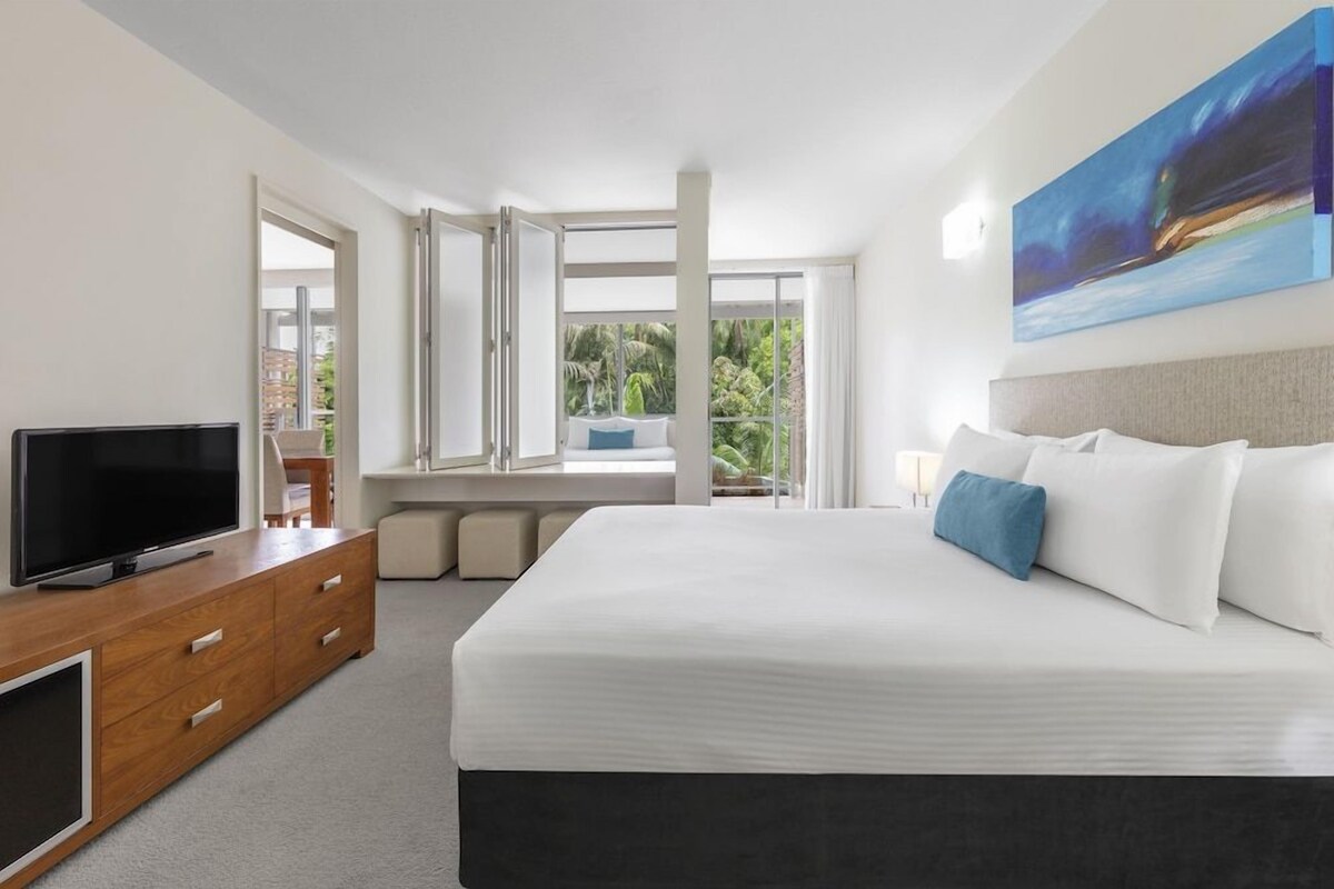 A modern hotel room with a large bed, a flat-screen TV, a wooden dresser, and a view of greenery through the windows.
