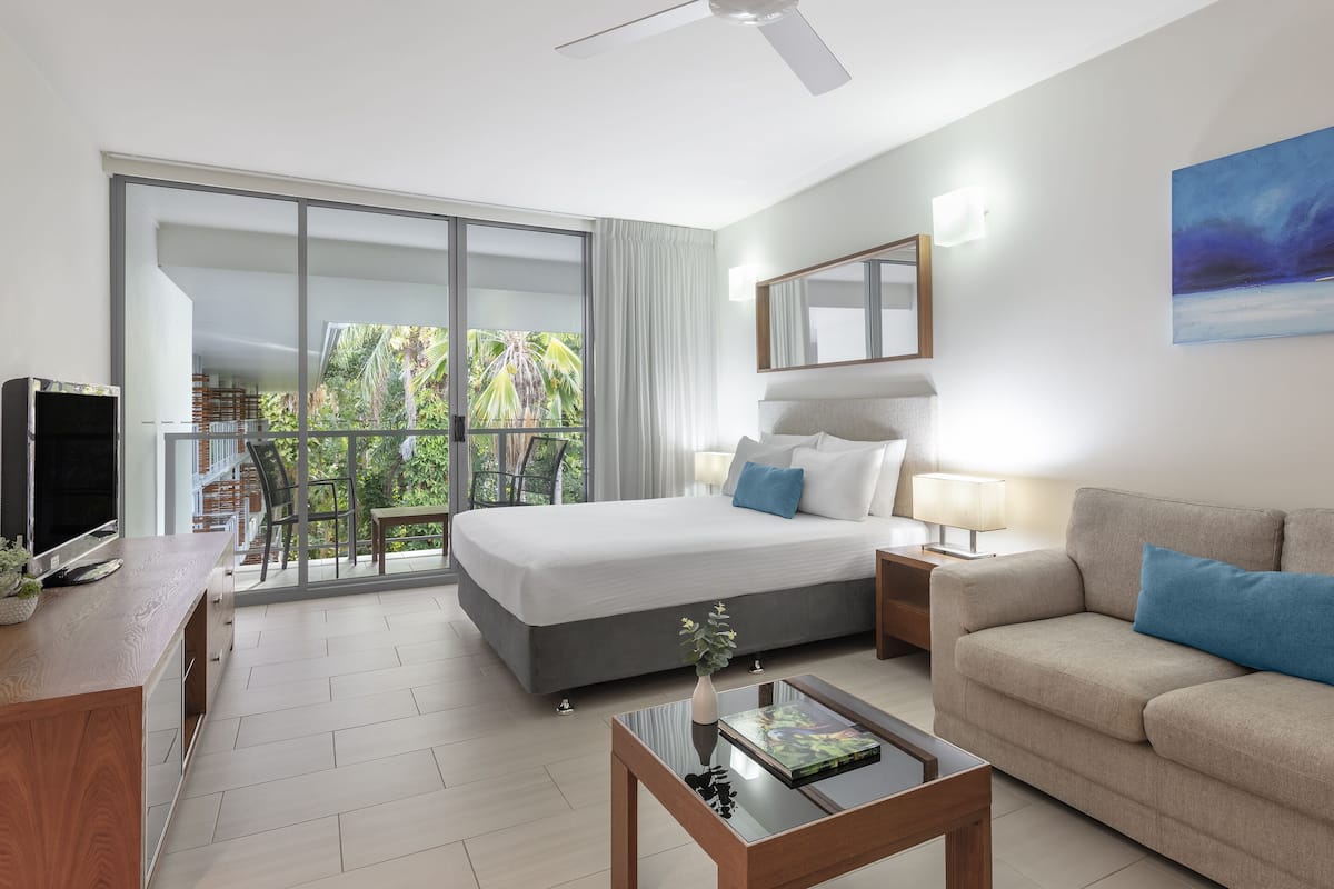 A modern hotel room with a large bed, a sofa, a coffee table, a TV, and a balcony with a view of palm trees.