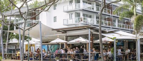Un immeuble à plusieurs étages doté d’une terrasse et de parasols, entouré de palmiers et de verdure.