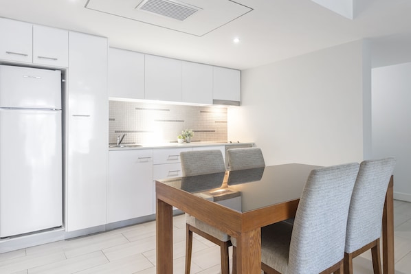 Una cocina moderna con armarios blancos, horno empotrado y comedor con mesa de vidrio y sillas tapizadas en gris.