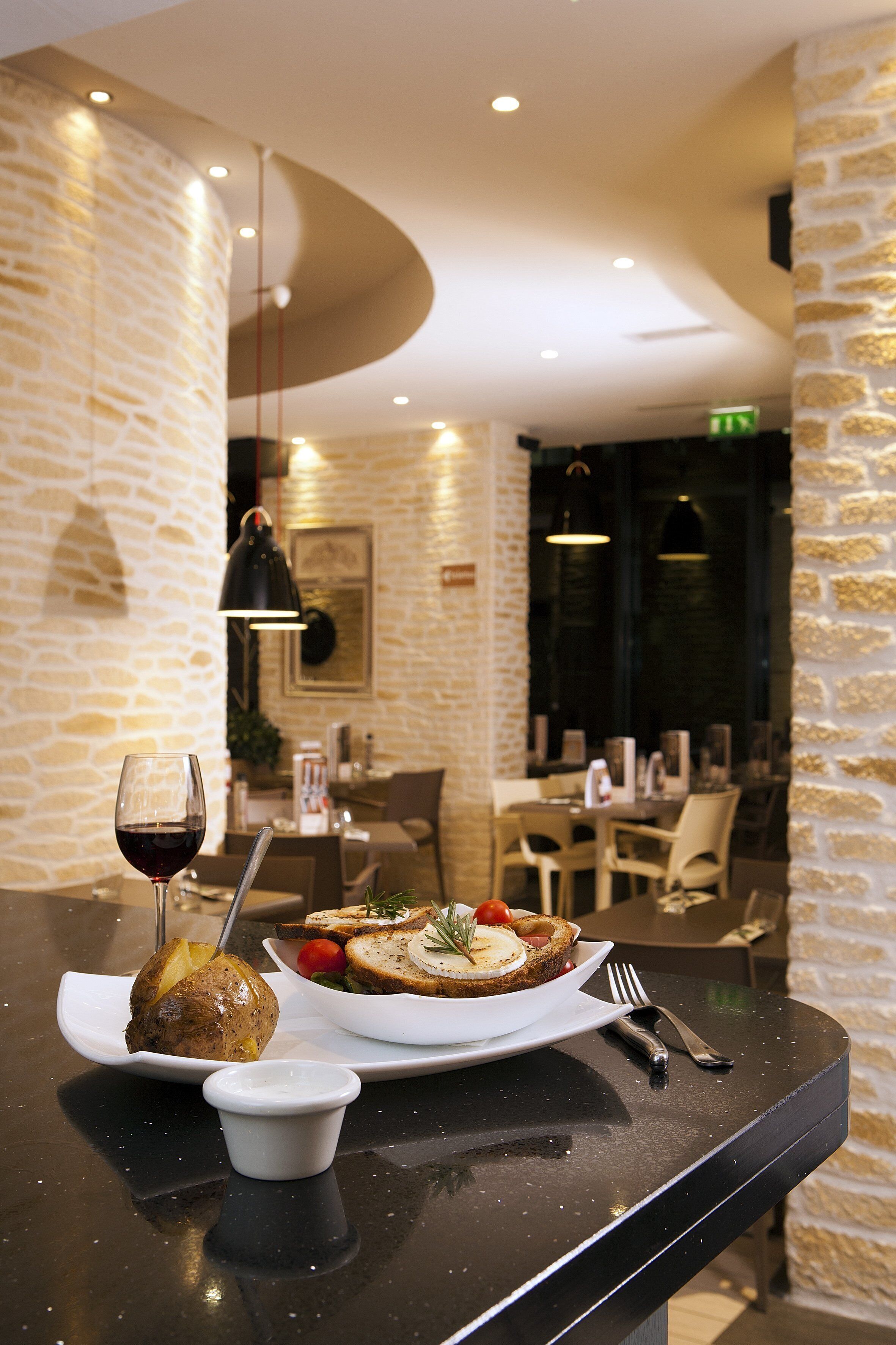 A dining table with a plate of food, a glass of red wine, and a small bowl of sauce. The setting includes stone walls and modern lighting fixtures.