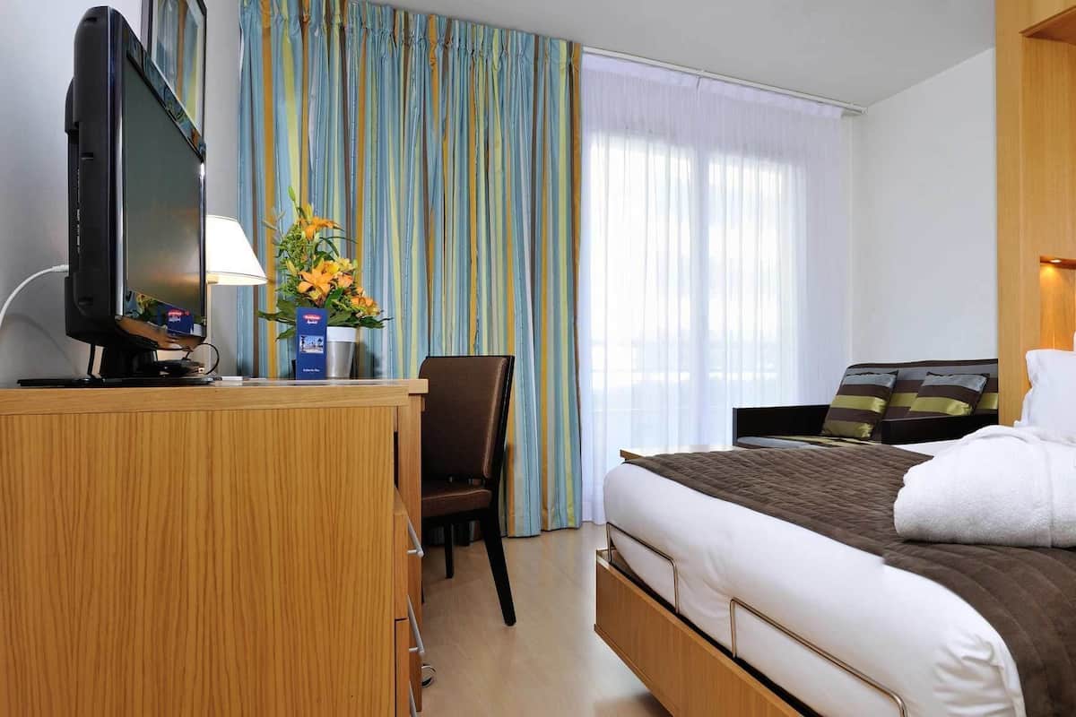 A hotel room with a bed, a desk with a computer, a chair, a TV, and a window with curtains.