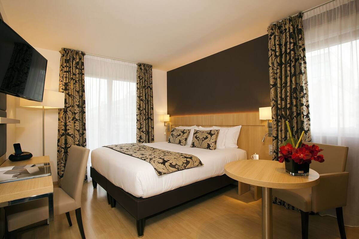 A hotel room with a bed, a desk, a chair, a TV, and a table with a vase of flowers.