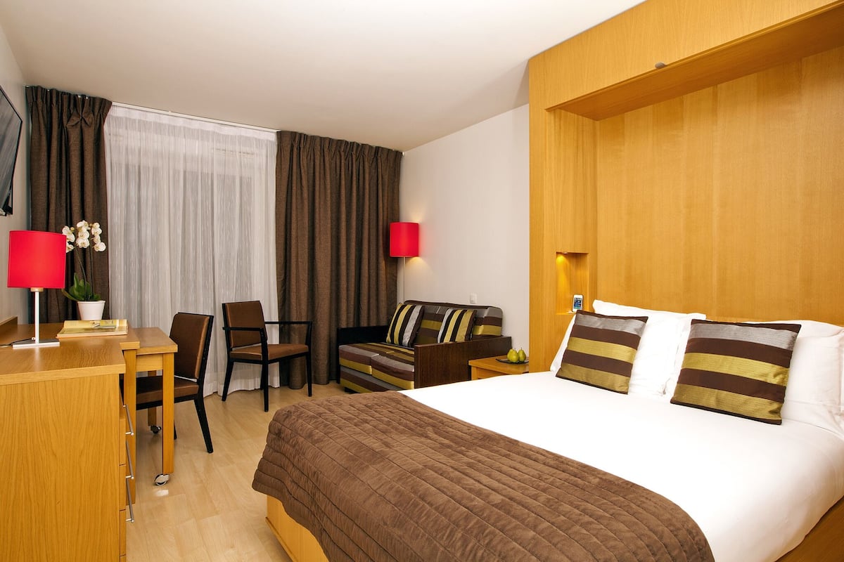 A hotel room with a bed, a desk, a chair, a sofa, and a small table with a lamp and a plant.