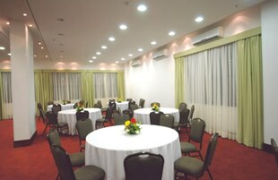 A conference room with multiple round tables, each with a white tablecloth and a flower centerpiece. The room has green chairs, a red carpet, and large windows with green curtains.