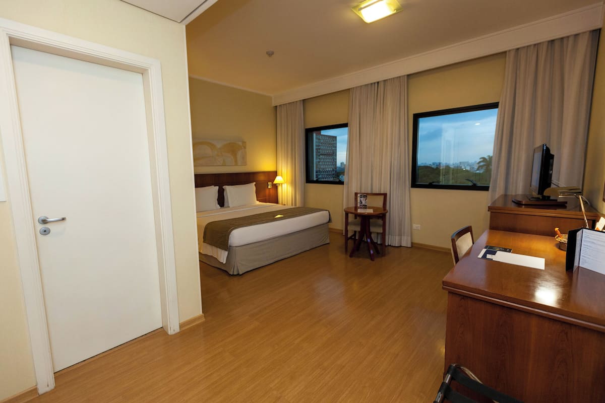 Une chambre d’hôtel avec un lit, un bureau, une chaise, une télévision et une fenêtre avec des rideaux.