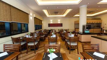 A spacious hotel dining area with multiple tables and chairs, a central column, and a kitchen area in the background.