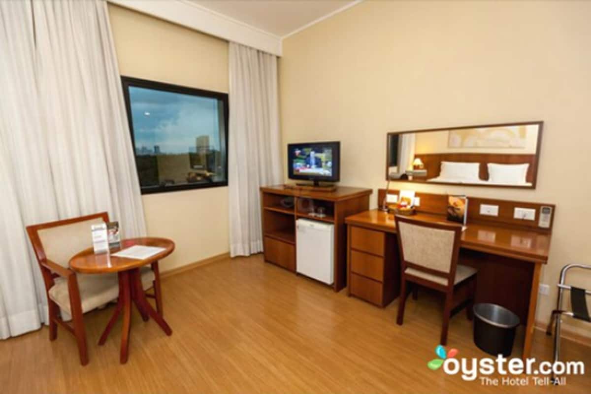 Une chambre d’hôtel équipée d’un bureau, de deux chaises, d’une télévision, d’un petit réfrigérateur, d’une poubelle et d’une fenêtre avec des rideaux.