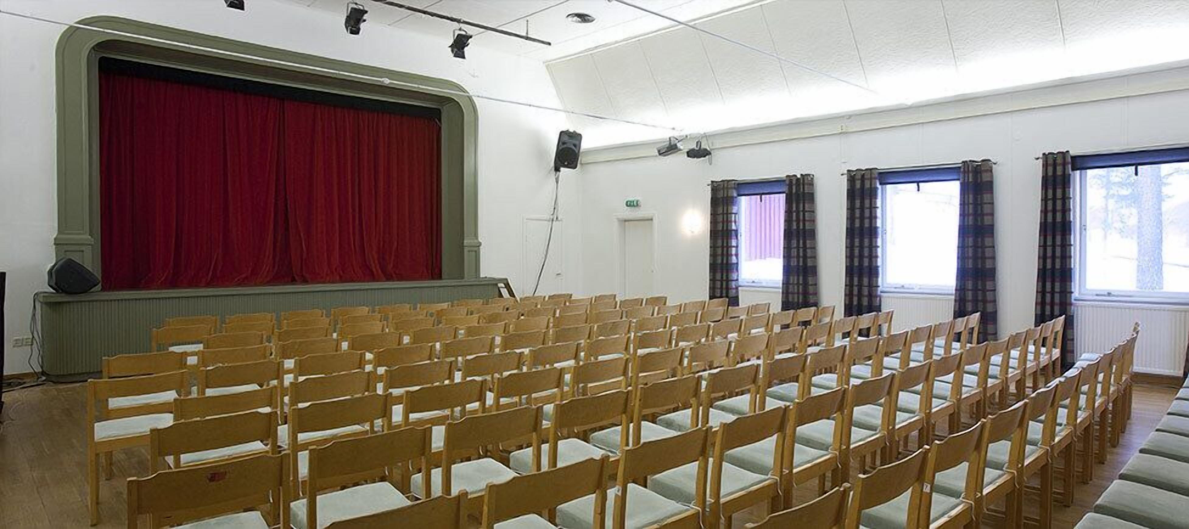 Un auditorio con filas de sillas de madera, un telón de fondo rojo y ventanas con cortinas de cuadros.
