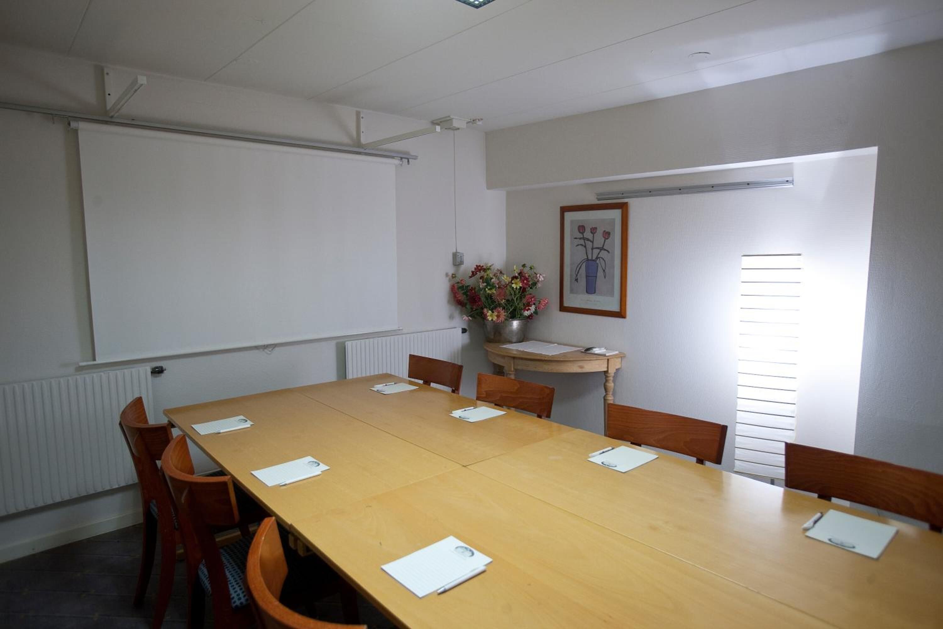 Un salón de conferencias con una larga mesa de madera, varias sillas, una pantalla de proyector, un cuadro enmarcado y una ventana con persianas.