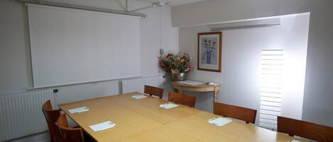 Un salón de conferencias con una larga mesa de madera, varias sillas, una pantalla de proyector, un cuadro enmarcado y una ventana con persianas.