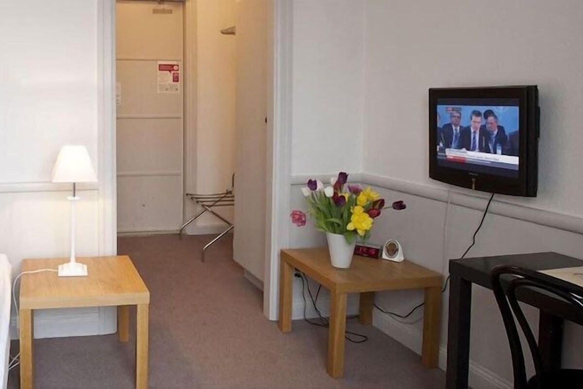 A hotel room with a bed, a bedside table with a lamp, a small table with a vase of flowers, a television on the wall, and a desk with a chair.