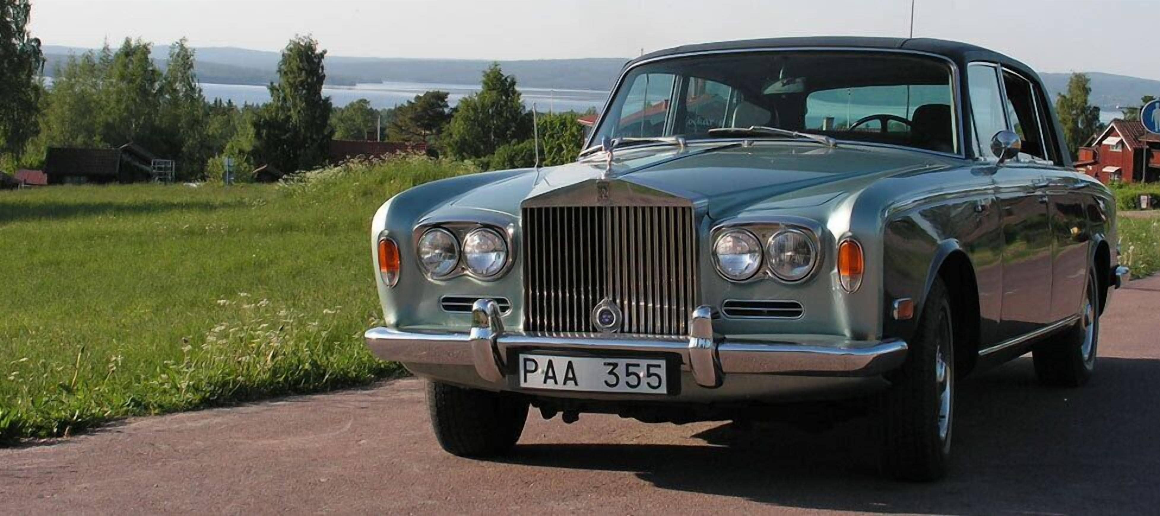 A vintage car parked on a paved road with a grassy field and trees in the background.
