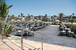A resort pool area with multiple lounge chairs and tables, surrounded by palm trees and umbrellas.